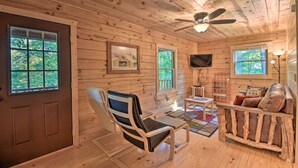 Interior de una cabaña de madera con ventilador de techo, televisión, sofá y sillón.