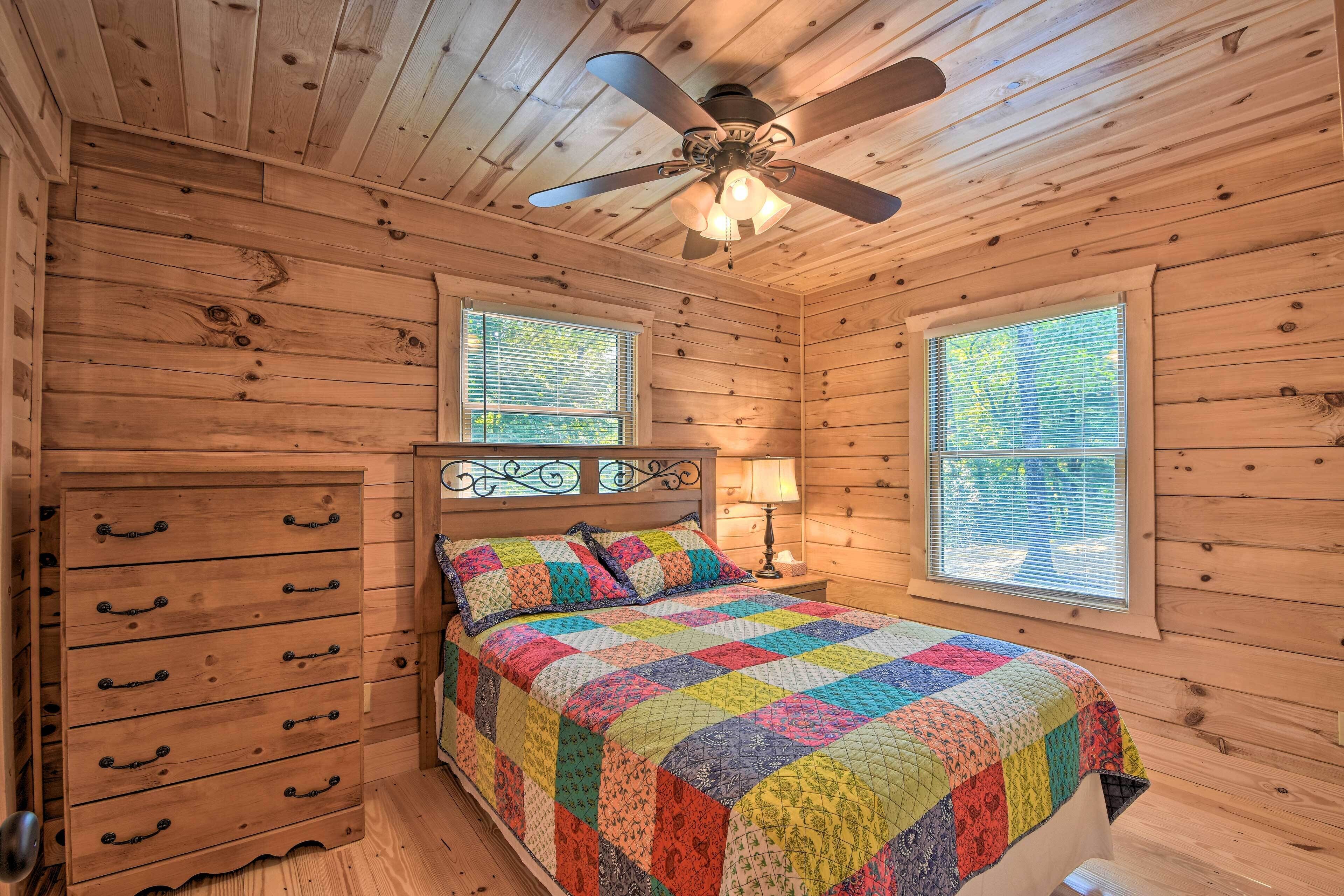 A wooden cabin bedroom with a bed, dresser, and two windows.