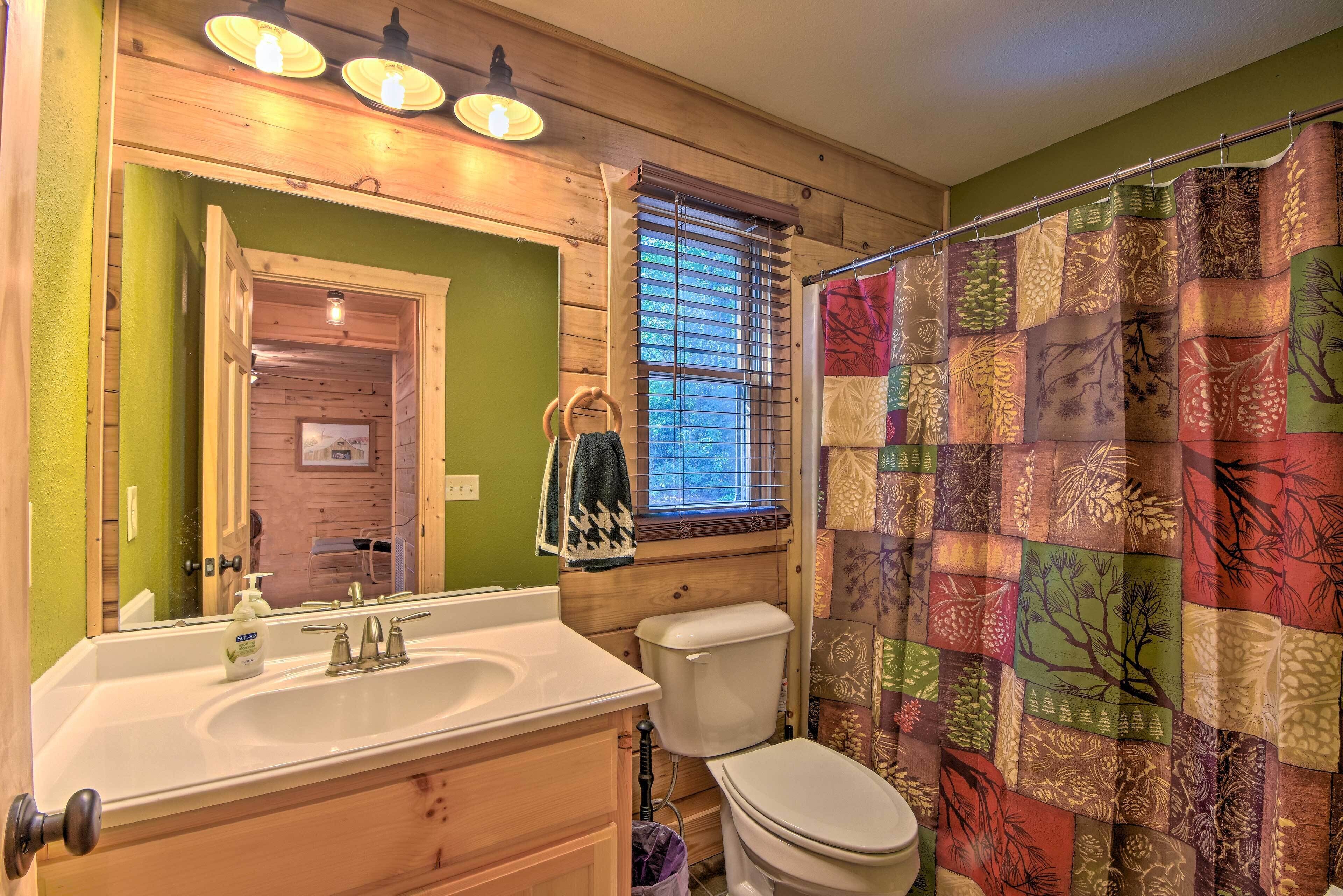 Un baño con tocador de madera, lavamanos blanco, inodoro y cortina de ducha con estampado de árboles.