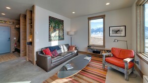 Un salon moderne avec un canapé en cuir, un fauteuil rouge et une table basse. On y trouve également une bibliothèque et une grande fenêtre donnant sur une vue imprenable sur les montagnes.