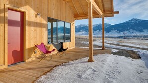 Une cabane en bois avec une porte rouge, deux chaises pliantes et une grande fenêtre donnant sur un paysage montagneux enneigé.