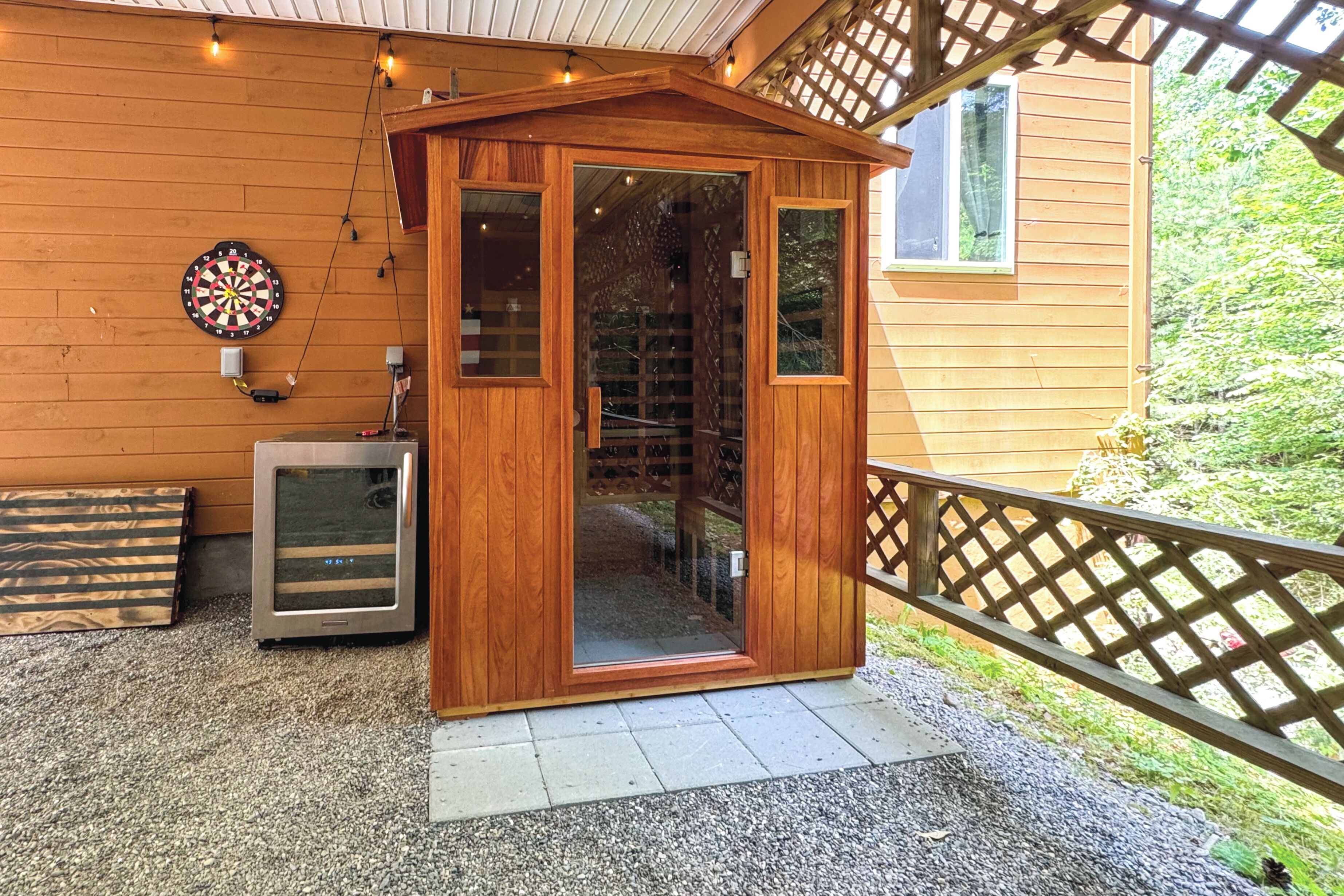 A wooden structure with a glass door, a dartboard, and a small refrigerator on a gravel surface.