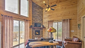 A cozy cabin interior with a fireplace, wooden floors, a ceiling fan, and a large window overlooking a deck. - Hot Tub & Views: Charming Bryson City Cabin! (Bryson City)