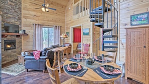 A cozy cabin interior with a fireplace, a dining table set for four, a sofa, and a spiral staircase. - Hot Tub & Views: Charming Bryson City Cabin! (Bryson City)