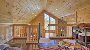 A wooden cabin interior with a loft area, wooden railings, and a view through a large window. - Hot Tub & Views: Charming Bryson City Cabin! (Bryson City)