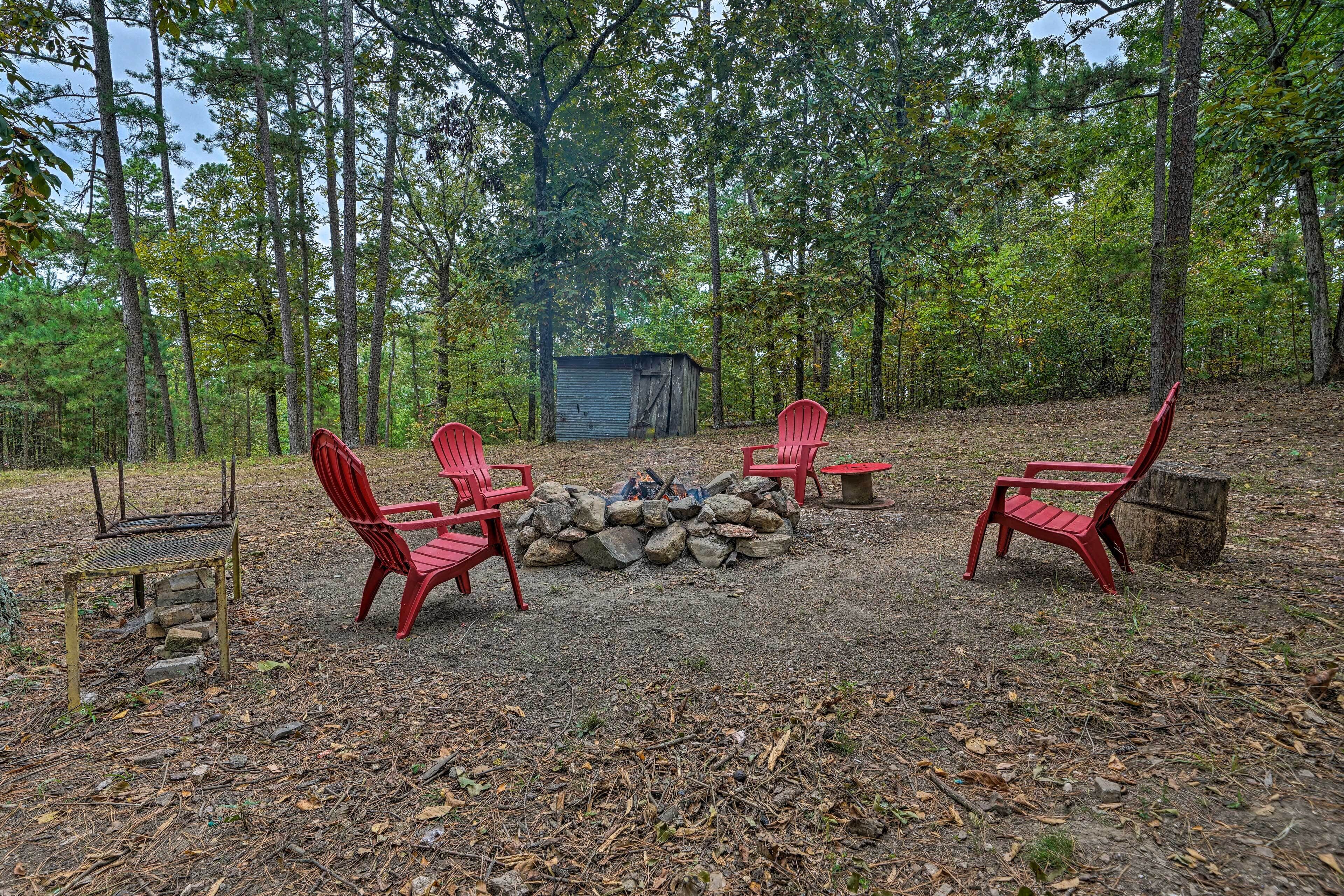 A rustic outdoor seating area with red chairs around a stone fire pit, surrounded by a forest.