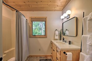 A bathroom with a wooden ceiling, a window with a view of trees, a sink with a mirror, and a towel rack.
