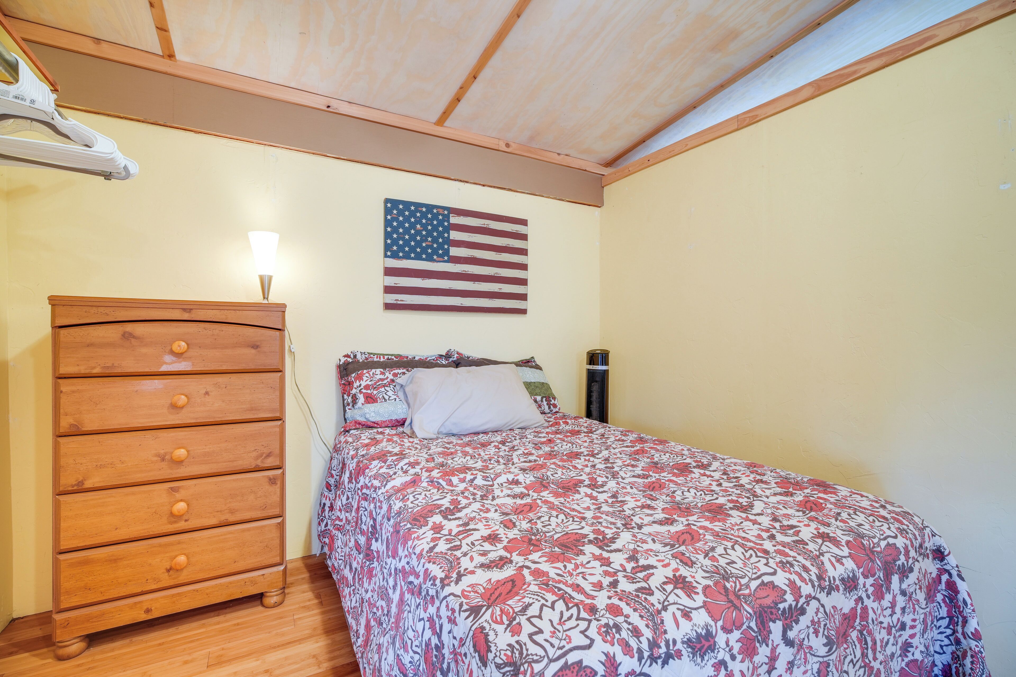 Een slaapkamer met een houten dressoir, een bed met een bloemenprint, een Amerikaanse vlag aan de muur en een plafondventilator.