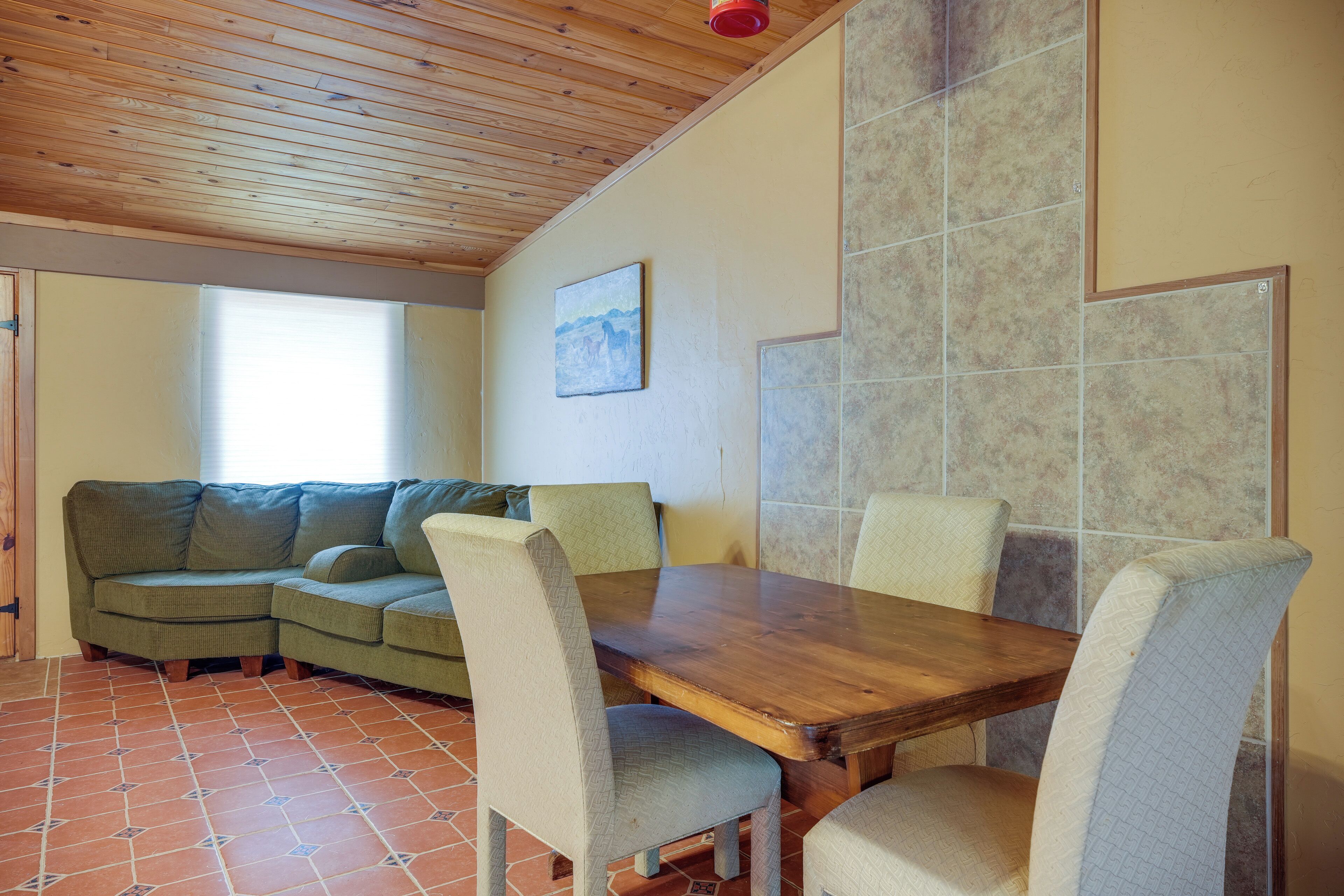 A cozy room with a wooden ceiling, a dining table with chairs, a green sofa, and a window with blinds.