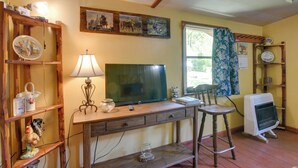 A room with a wooden desk, a television, a lamp, a chair, and a heater. - Serene Green Forest Hideaway w/ Spacious Yard (Green Forest)