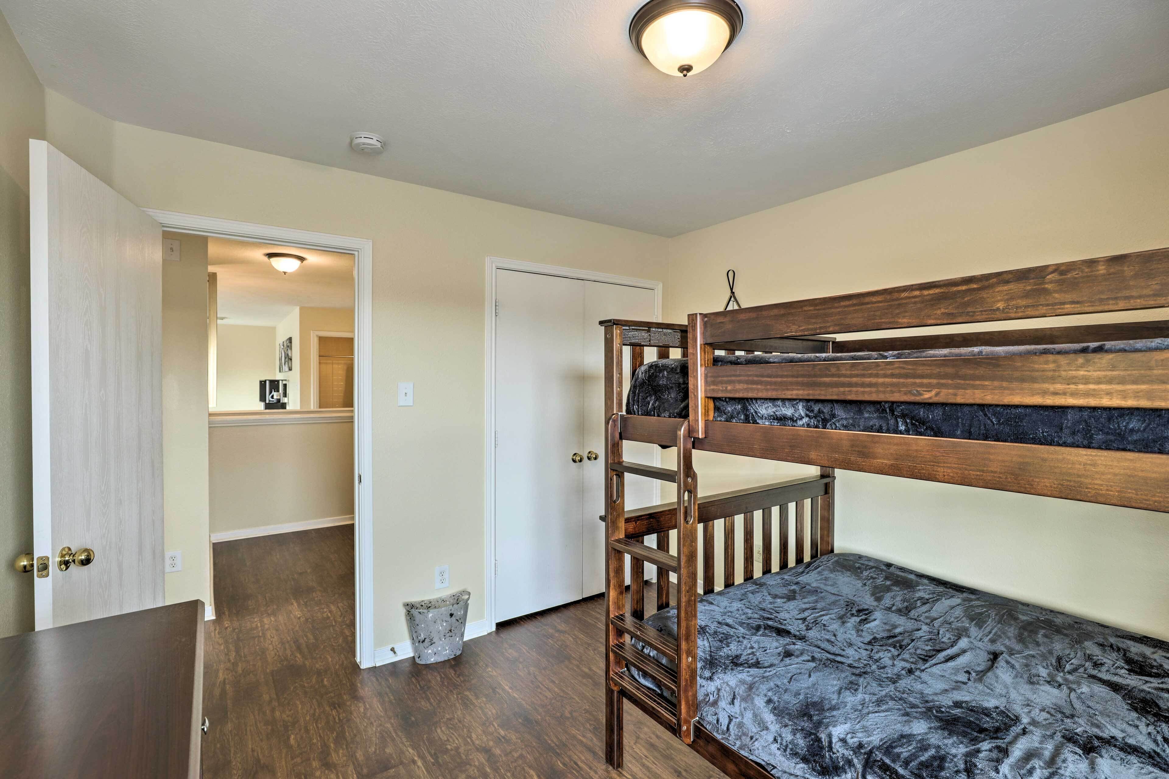 A bunk bed with a dark mattress, a wooden headboard, and a small ladder. There is a door leading to another room, a light fixture on the ceiling, and a trash can near the door.