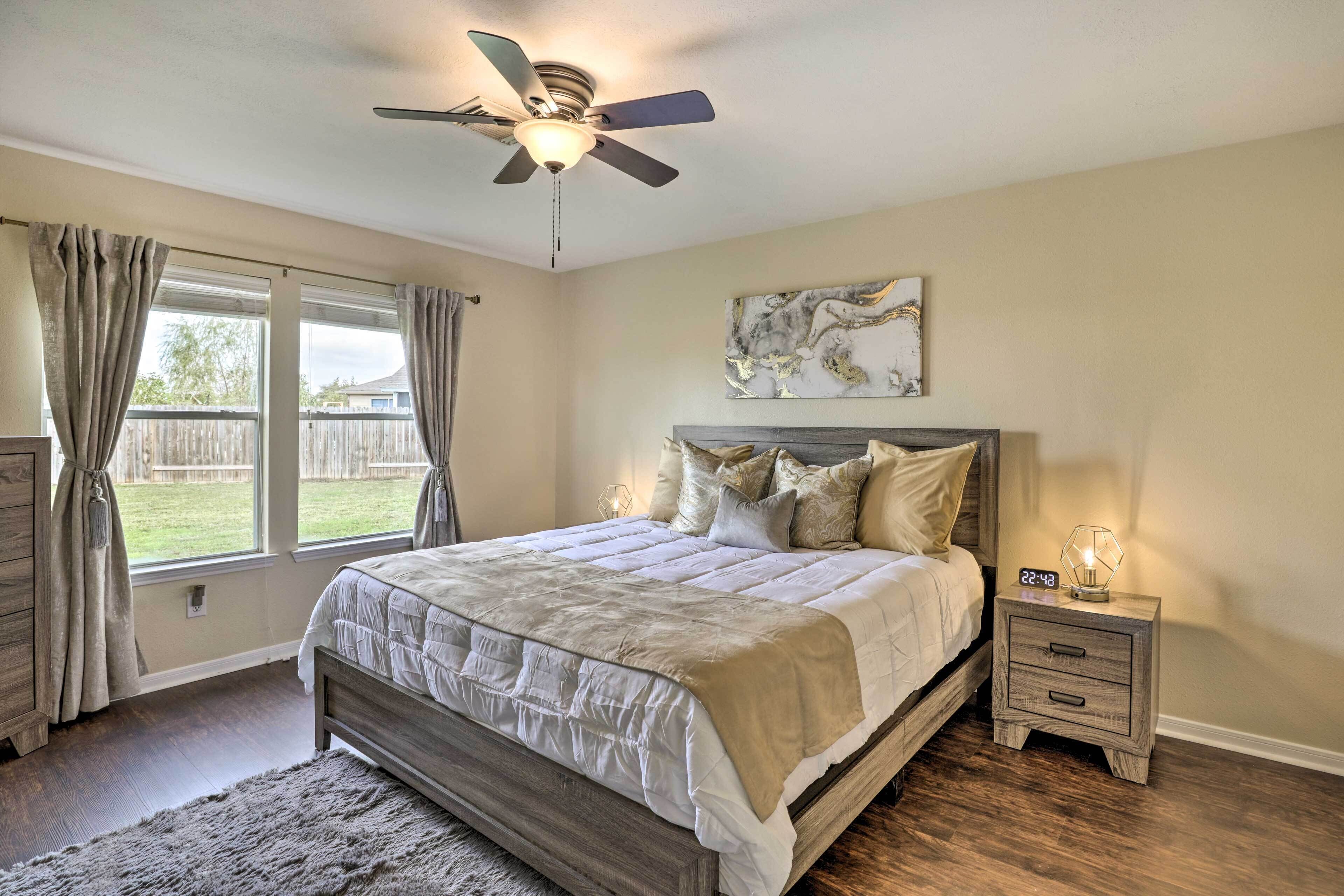 A bedroom with a bed, a nightstand, a ceiling fan, a window with curtains, and a wall-mounted artwork.