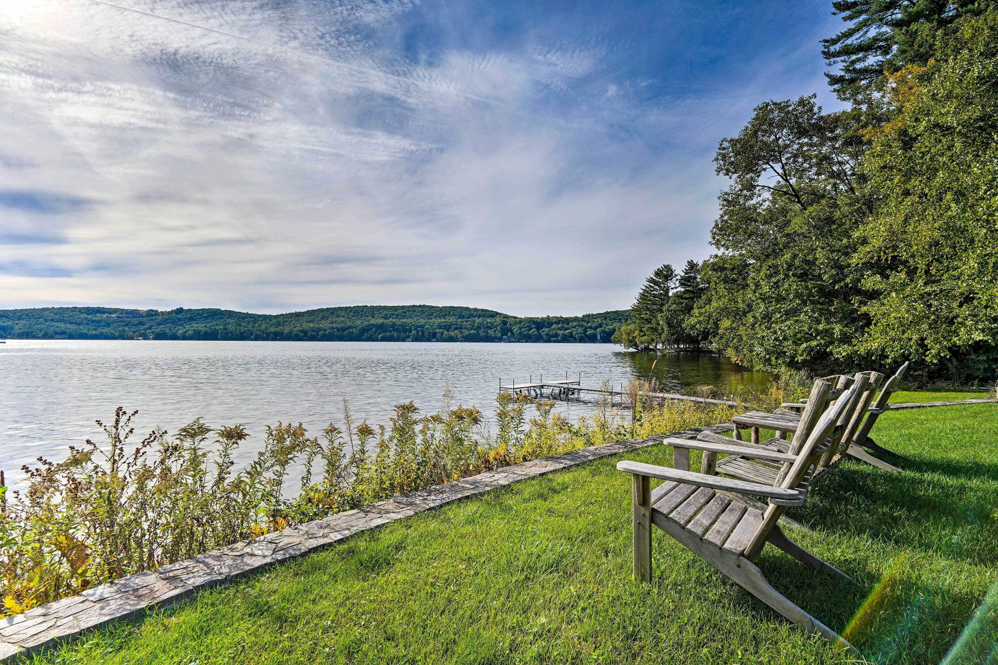 A lakeside view with a wooden dock and chairs on the grass.