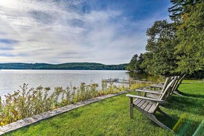 Blick auf einen See mit einer Holzsteg und Stühlen am Rasen.