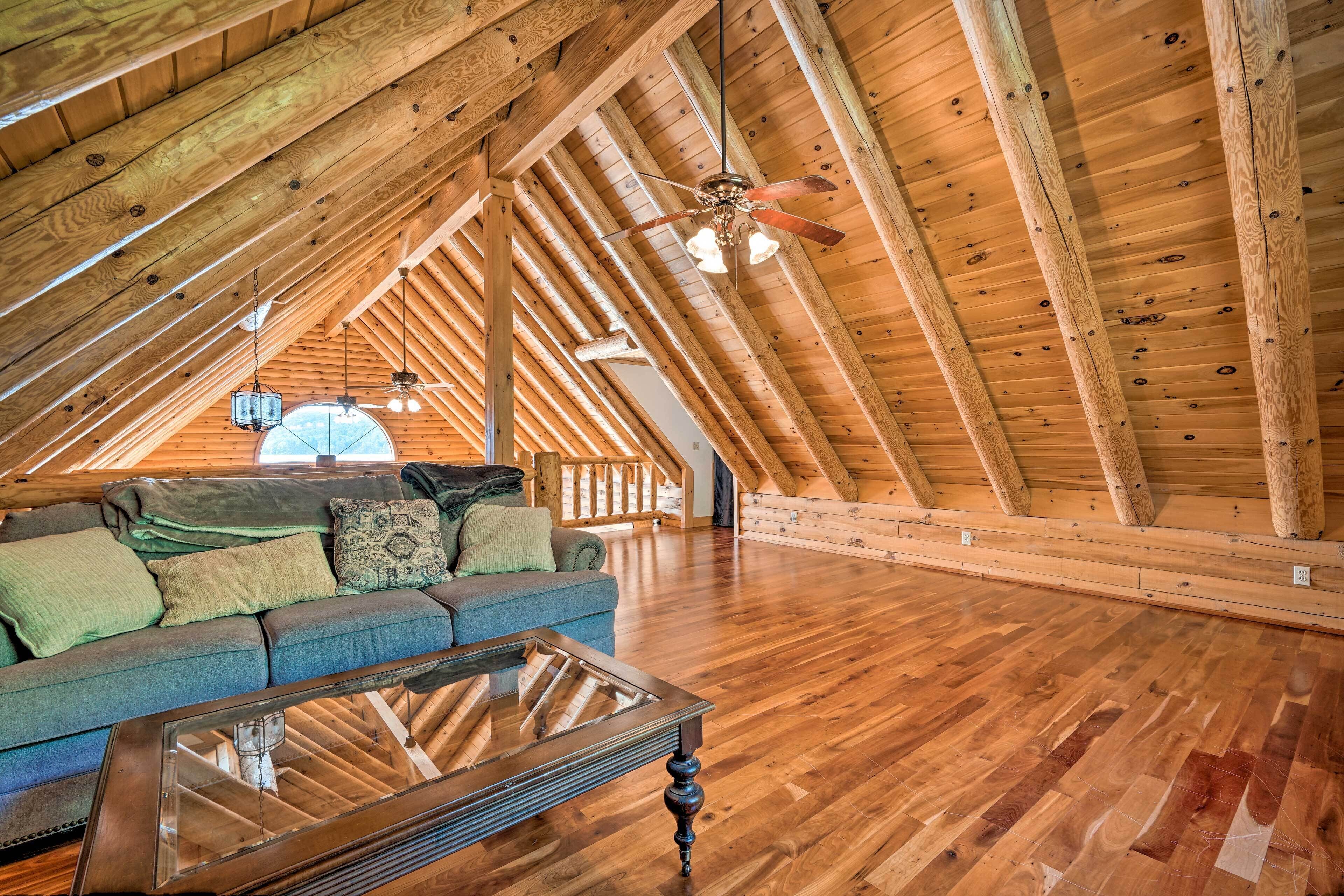 A spacious attic room with wooden beams, a glass coffee table, a sofa with cushions, and a ceiling fan.