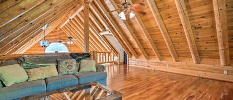 A spacious attic room with wooden beams, a glass coffee table, a sofa with cushions, and a ceiling fan.
