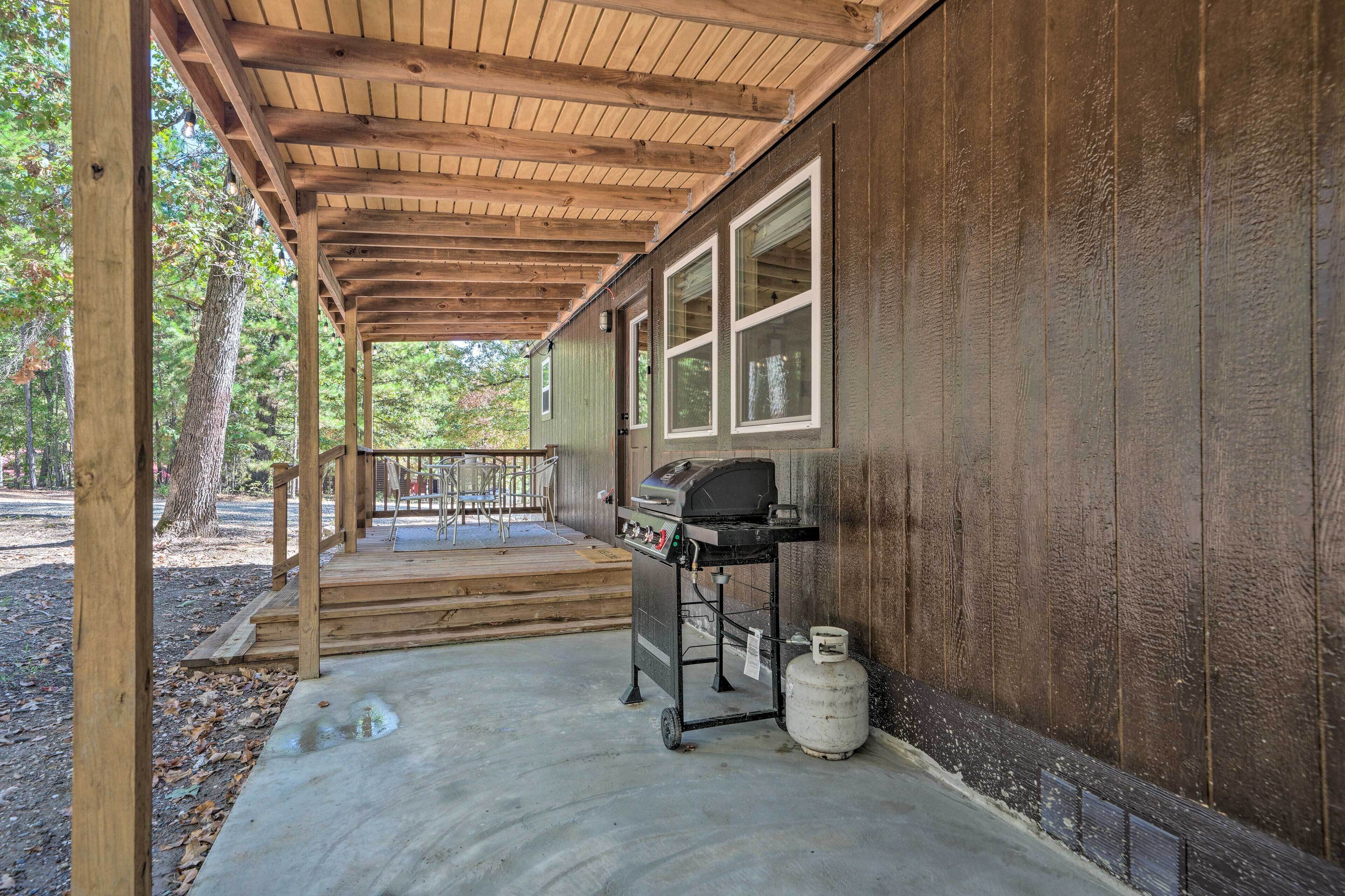 A covered porch with a grill and propane tank, wooden steps leading to a deck, and a view of a wooded area.
