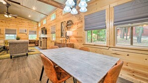 A spacious wooden cabin interior with a dining table, chairs, a ceiling fan, and large windows. - Stylish Cabin by Beavers Bend: Hot Tub & Fire Pit (Broken Bow)