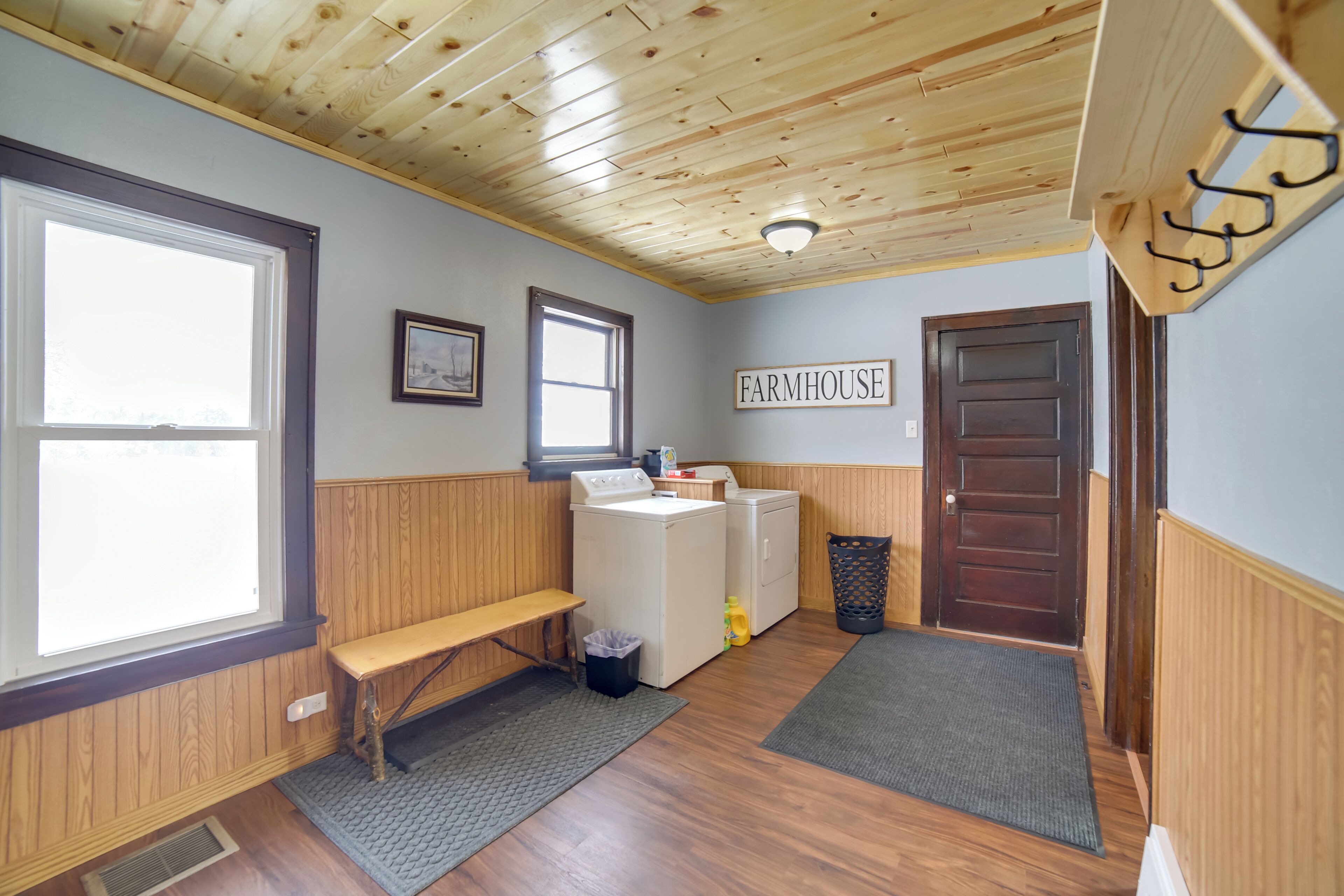 A laundry room with wooden walls, a bench, a washer and dryer, a trash can, and a door labeled "FARMHOUSE".