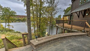 Une terrasse en bois avec une balustrade en pierre donnant sur un lac, une maison avec un toit en métal et des arbres aux feuilles d’automne.