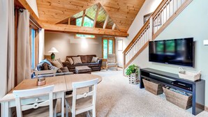 Un salon chaleureux avec un plafond en bois, un téléviseur posé sur un meuble noir, un canapé, une table à manger avec des chaises et un escalier menant à l’étage supérieur.
