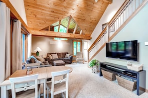 A spacious living room with a high ceiling, wooden beams, a large TV, a dining area, and a staircase.