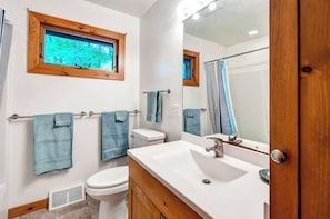 Une salle de bain avec un lavabo blanc, un meuble-lavabo en bois, des toilettes et une fenêtre donnant sur des arbres.
