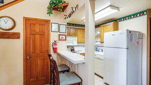 A kitchen with wooden cabinets, a white refrigerator, a wooden door, a fire extinguisher, and a clock on the wall. - Warm Mccall Condo: Half-mile to Payette Lake! (McCall)