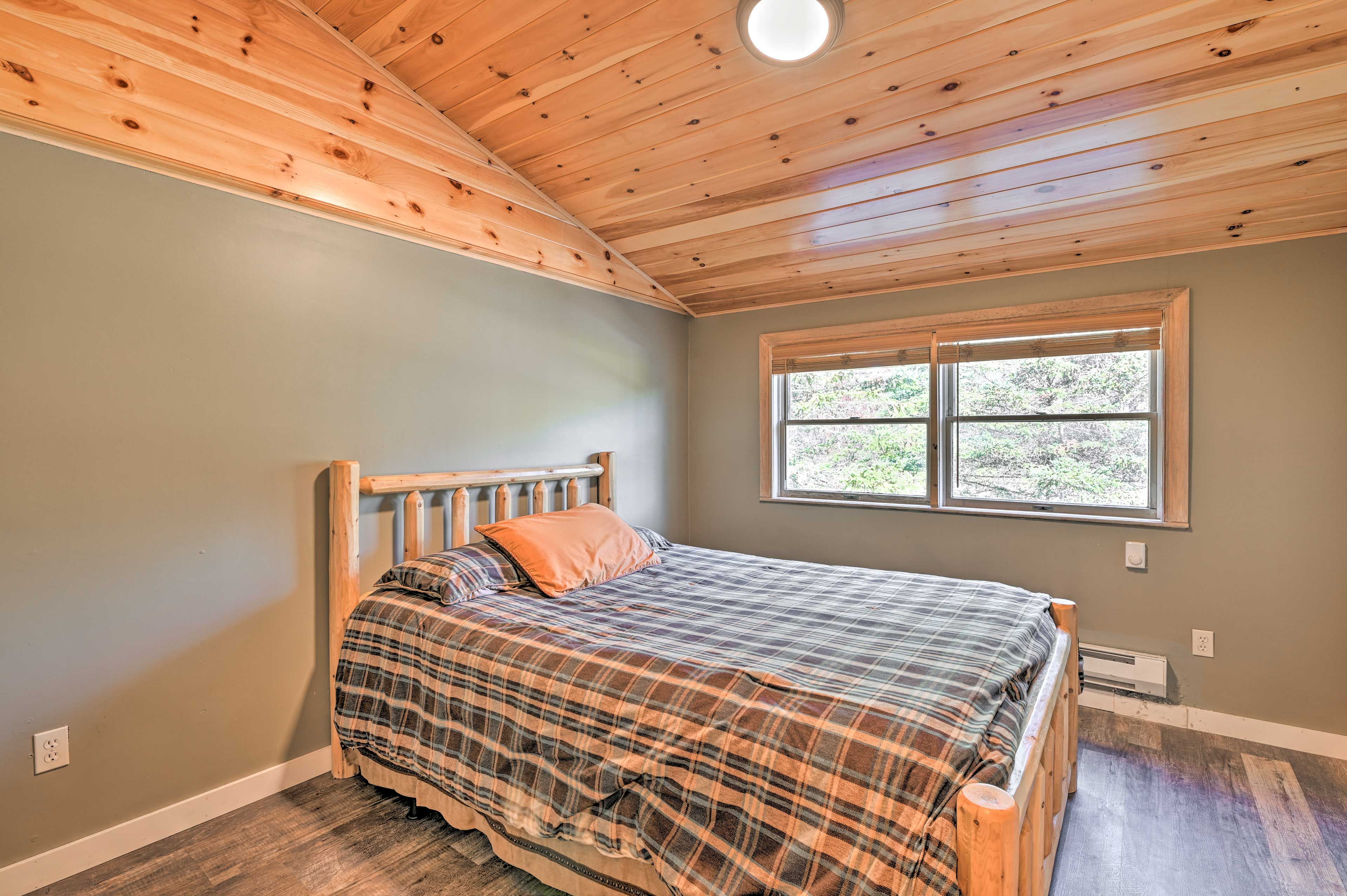 A bedroom with a wooden ceiling, a bed with a plaid bedspread, an orange pillow, and a window with a view of trees.