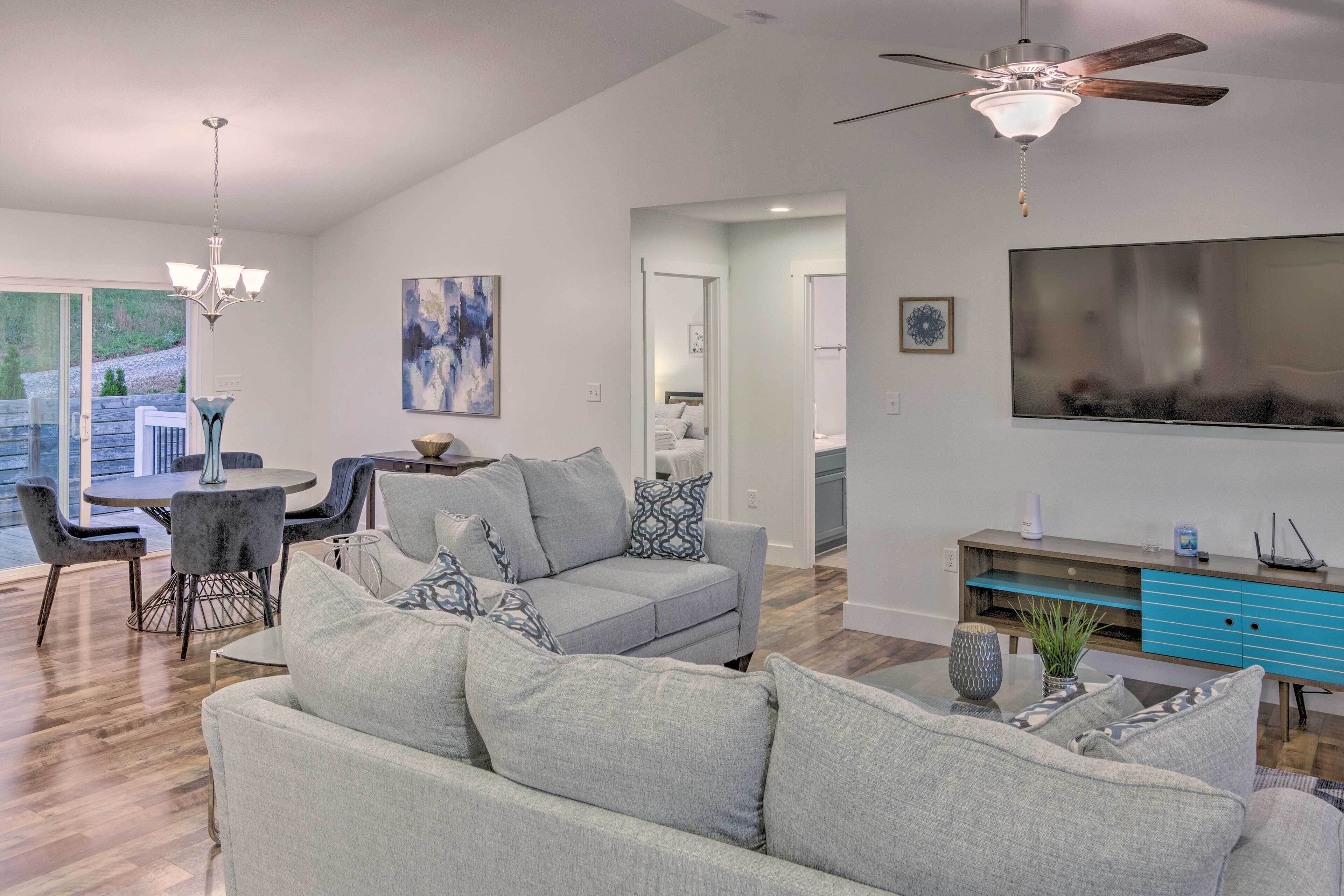 A modern living room with a grey sofa, a dining table with chairs, a ceiling fan, and a TV.