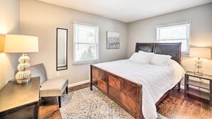 Une chambre avec un grand lit, deux tables de chevet, une chaise et un bureau. Il y a deux fenêtres avec des stores, un tableau au mur et un tapis à motifs sur le sol.