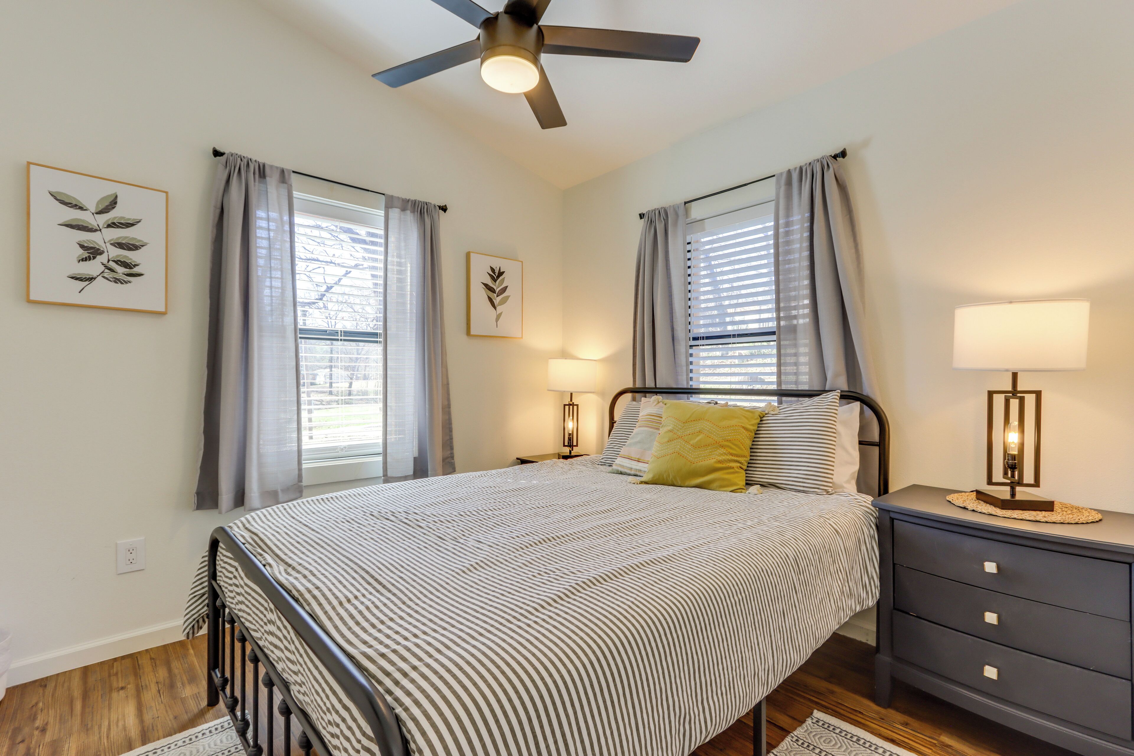 A bedroom with a bed, bedside tables, a ceiling fan, and two windows with curtains.