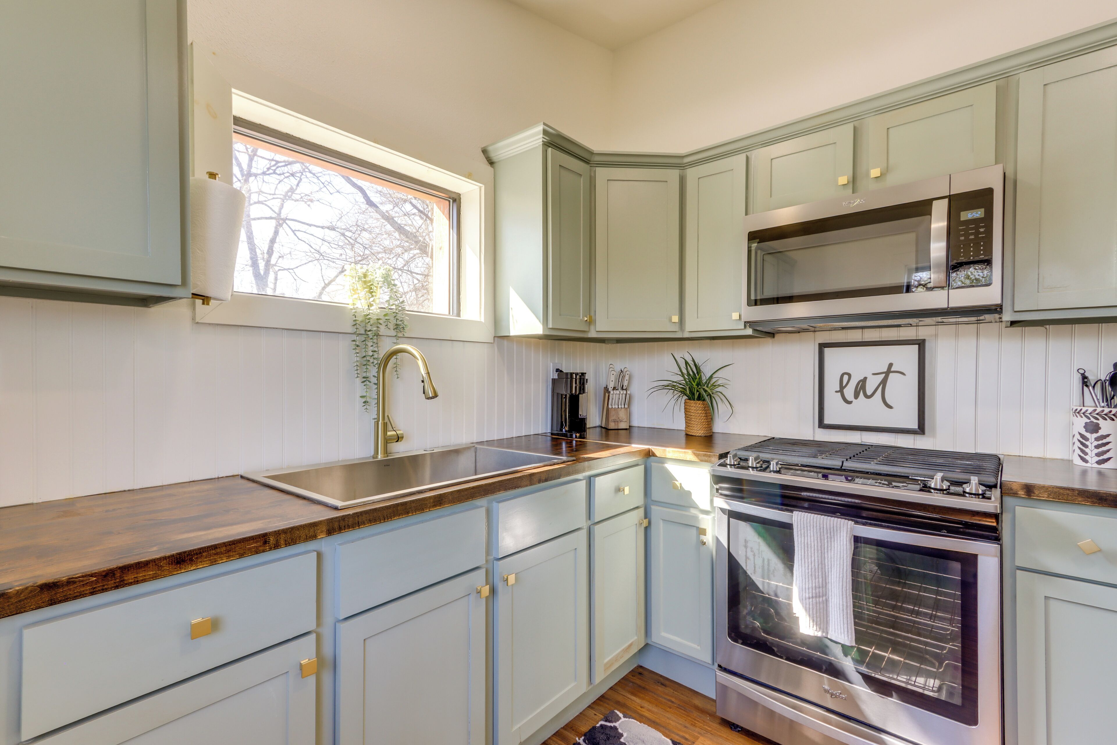 A kitchen with light blue cabinets, a stainless steel oven, a microwave, and a window with a plant.