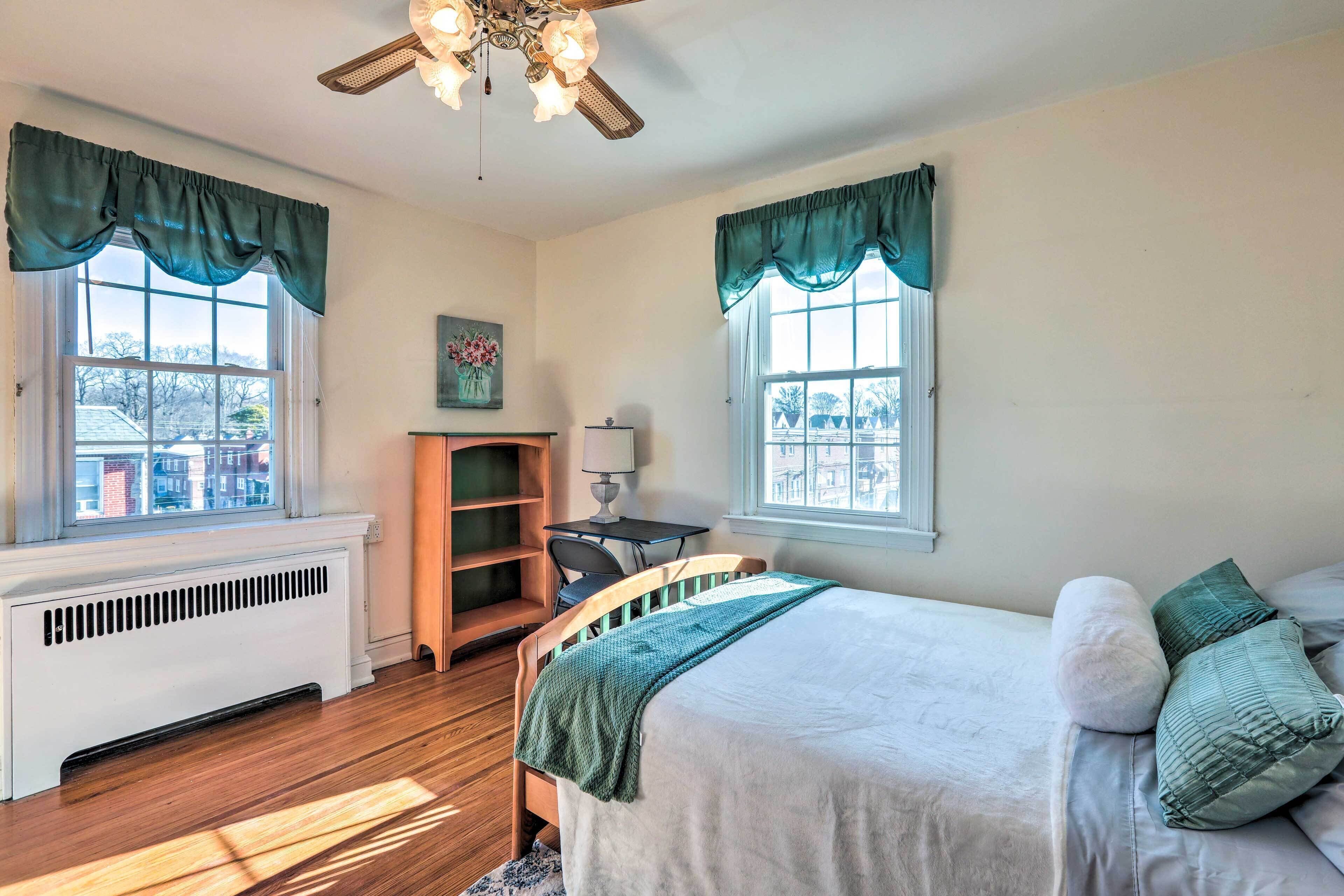 Un dormitorio con cama, escritorio, silla, radiador y ventanas con cortinas.