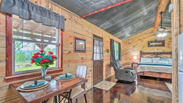 A wooden cabin interior with a dining table, chairs, a bed, and a ceiling fan. - Updated Studio Cabin in Ozark w/ Yard & Mtn View (Ozark)
