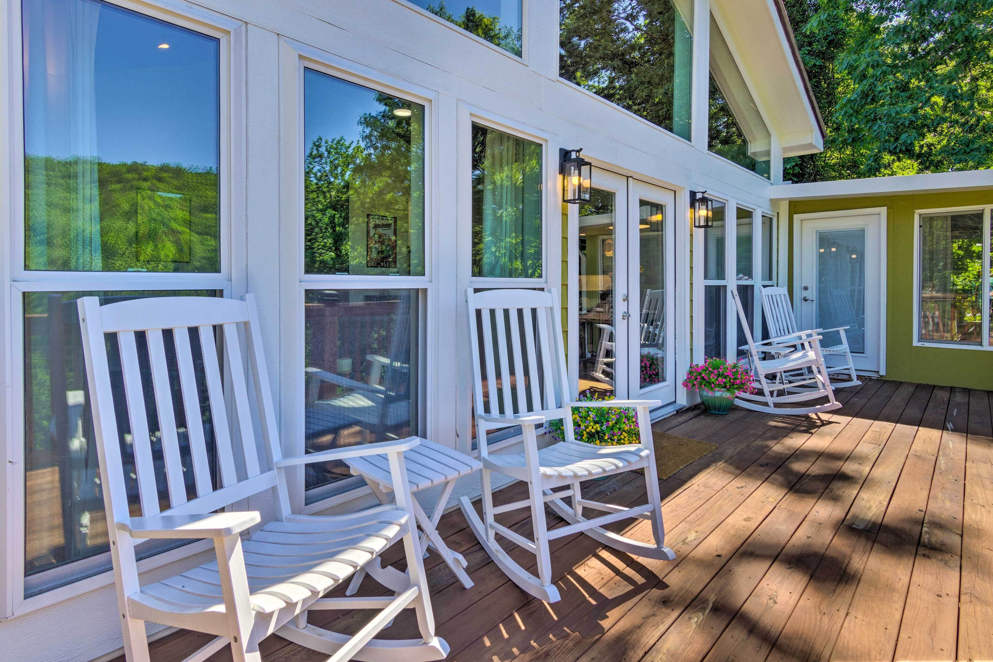 A wooden deck with white rocking chairs and large windows overlooking a garden.