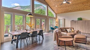 Una habitación amplia con techo alto, ventanas grandes que ofrecen vistas a un paisaje montañoso, un comedor con mesa y sillas, y una zona de estar acogedora con un sofá de cuero y reposapiés.