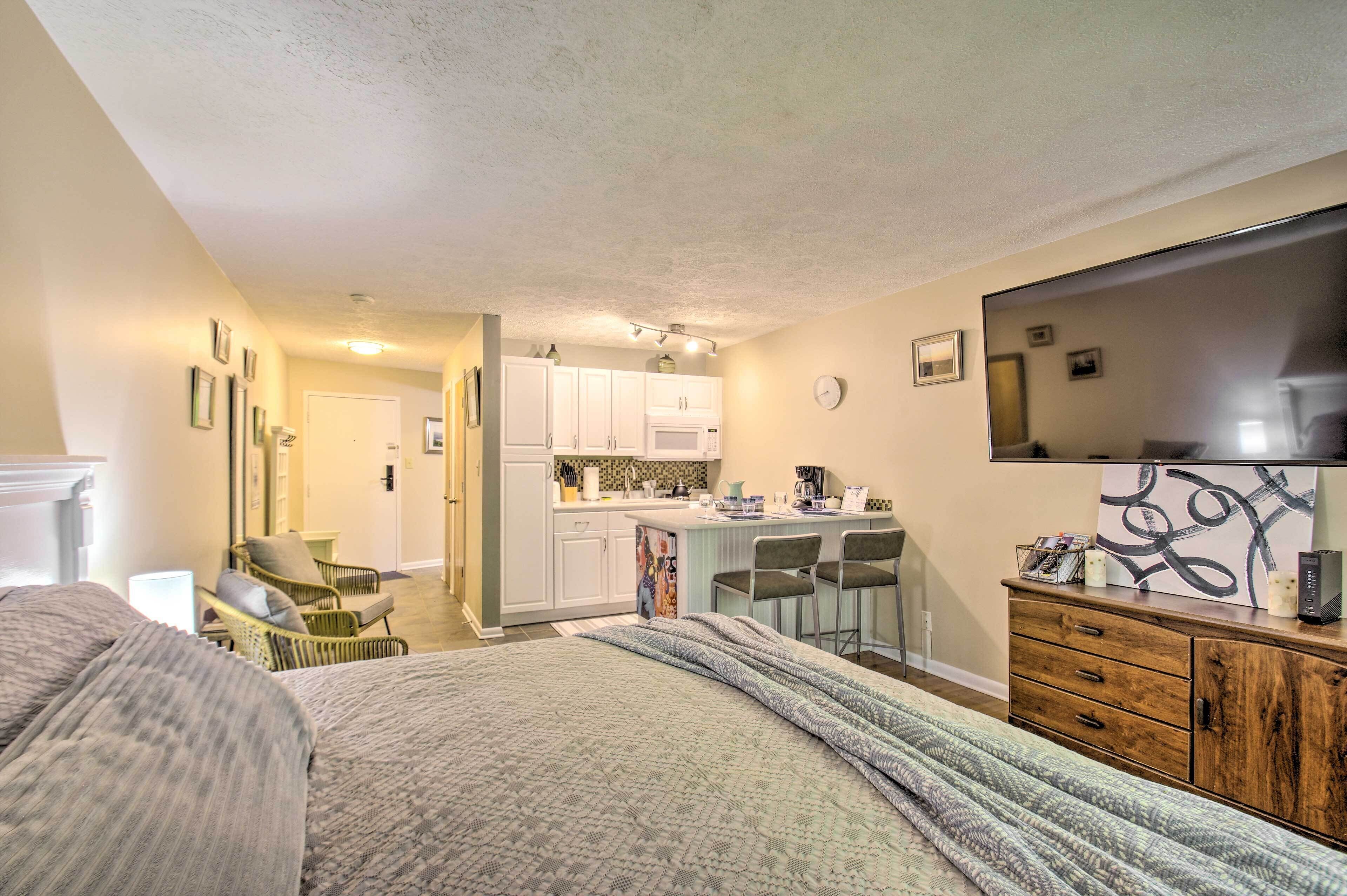 A compact hotel room with a large bed, a kitchenette, a dining area, and a TV mounted on the wall.