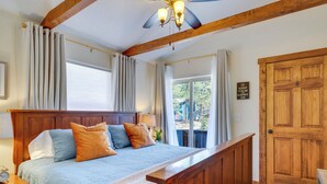 A bedroom with a wooden headboard, a large bed with patterned bedding, a nightstand with a clock, and a view of the outdoors through a window. - Woodland Park Apartment: Forest Views + Deck! (Woodland Park)