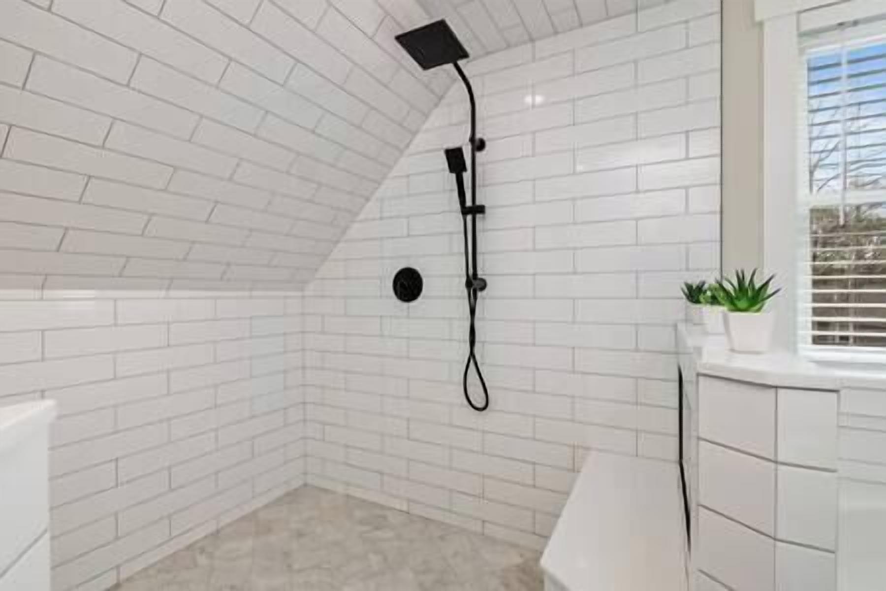 A small, white-tiled bathroom with a shower, a round mirror, and a potted plant.