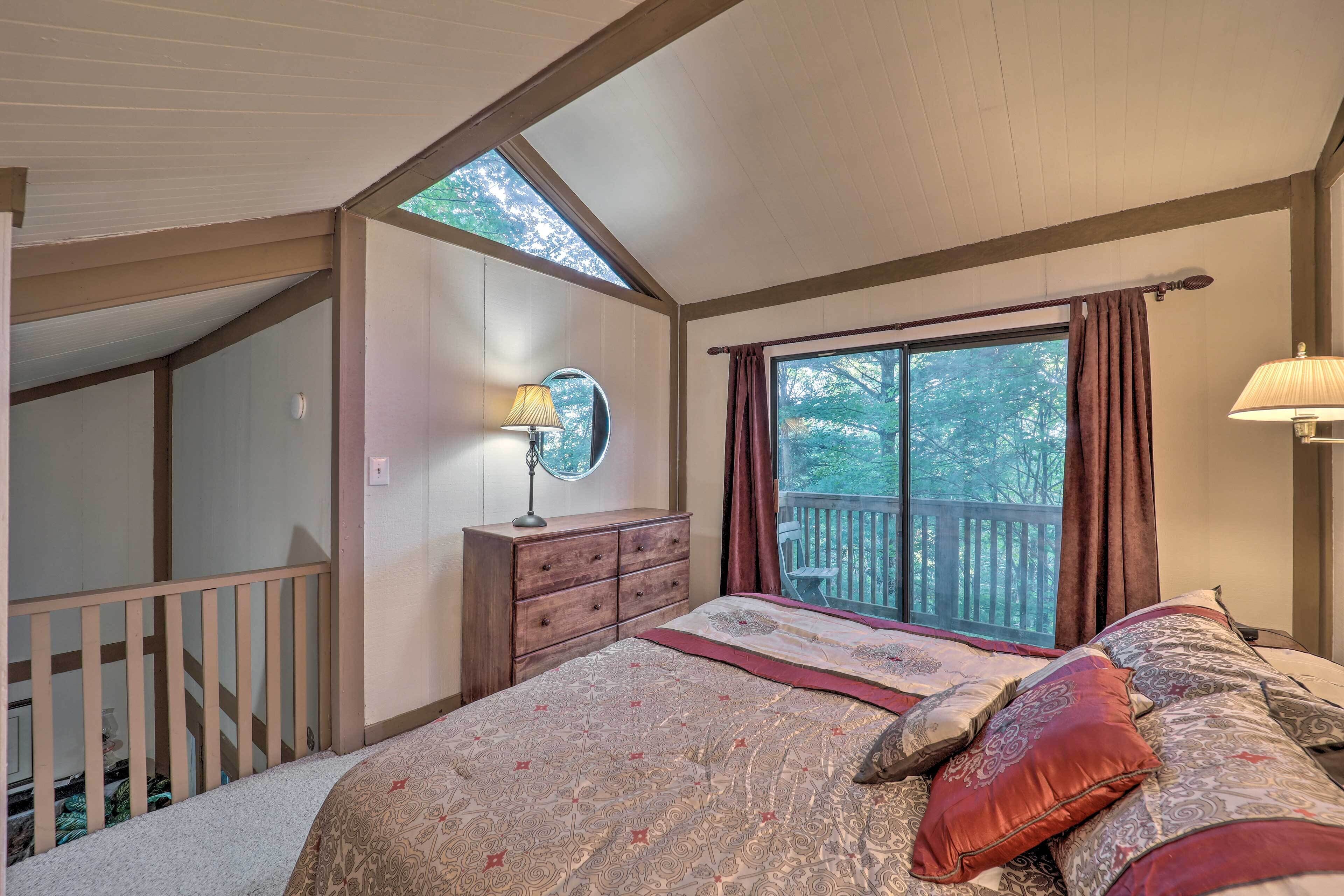 A bedroom with a large bed, a wooden dresser, a nightstand with a lamp, a window with curtains, and a skylight.