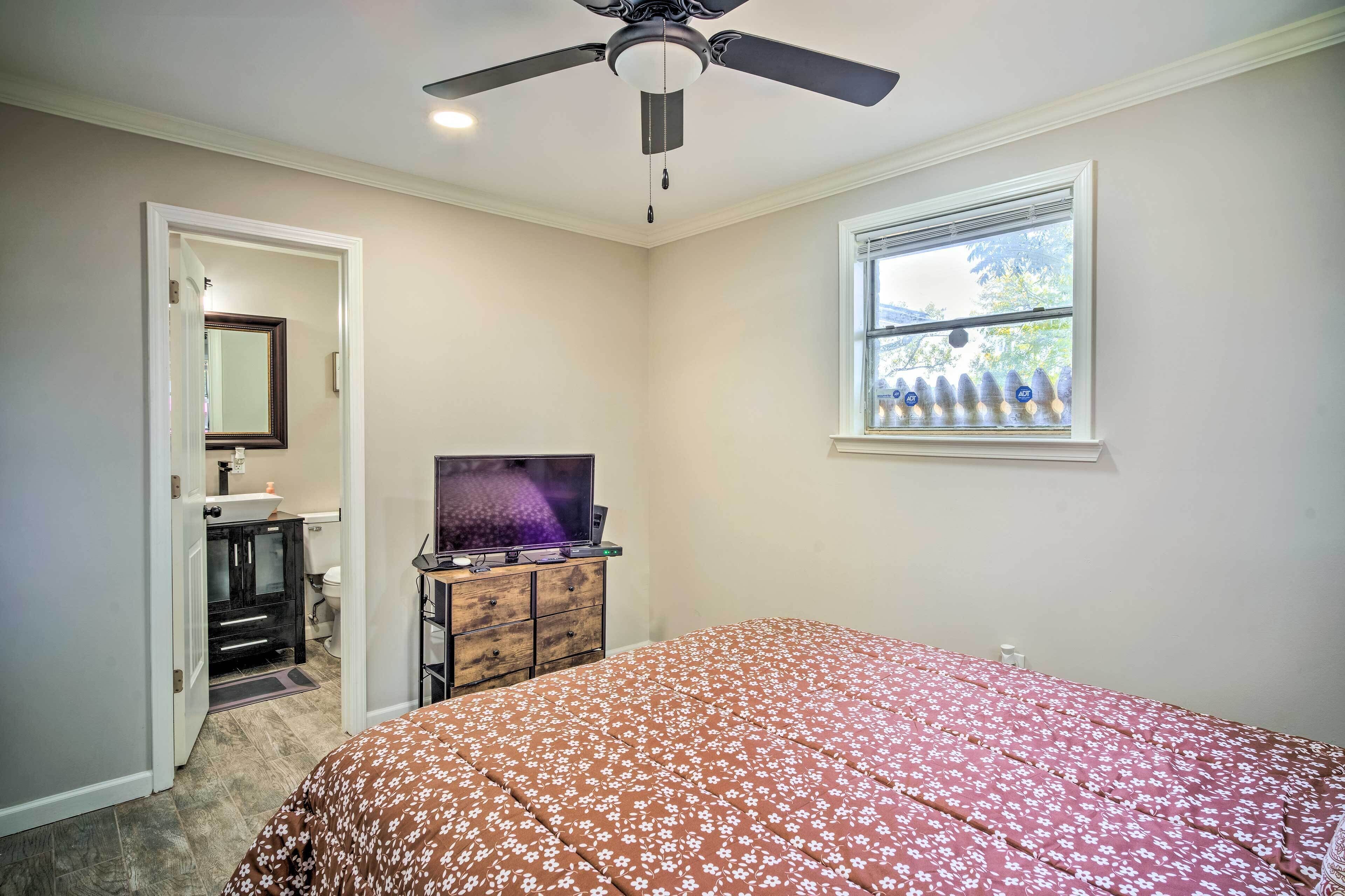 Une chambre à coucher comprenant un lit, un ventilateur de plafond, un téléviseur posé sur une commode, une fenêtre avec des stores et une porte donnant sur une autre pièce.