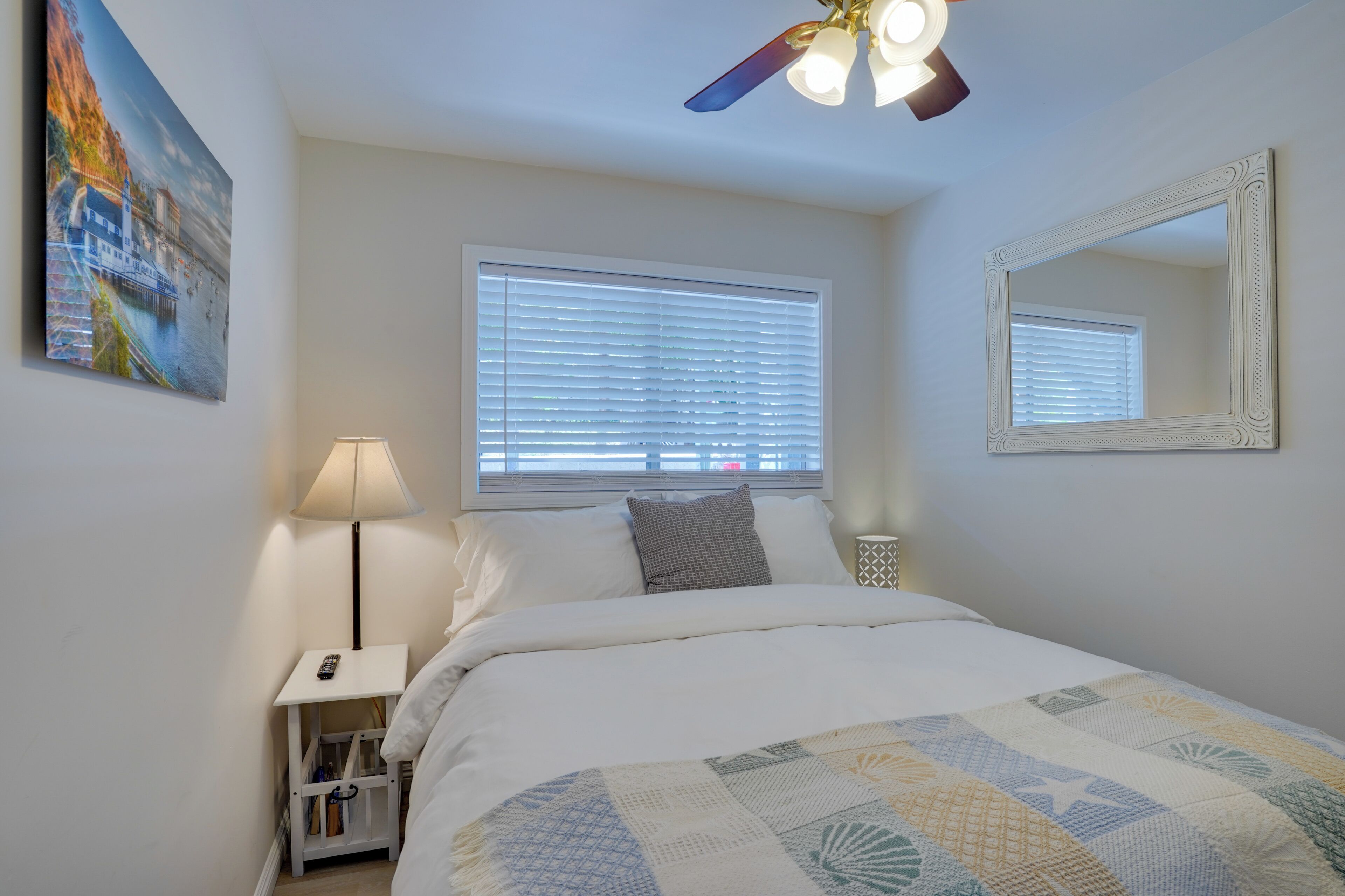 Un dormitorio con cama, mesita de noche, lámpara, ventilador de techo, ventana con persianas y espejo.