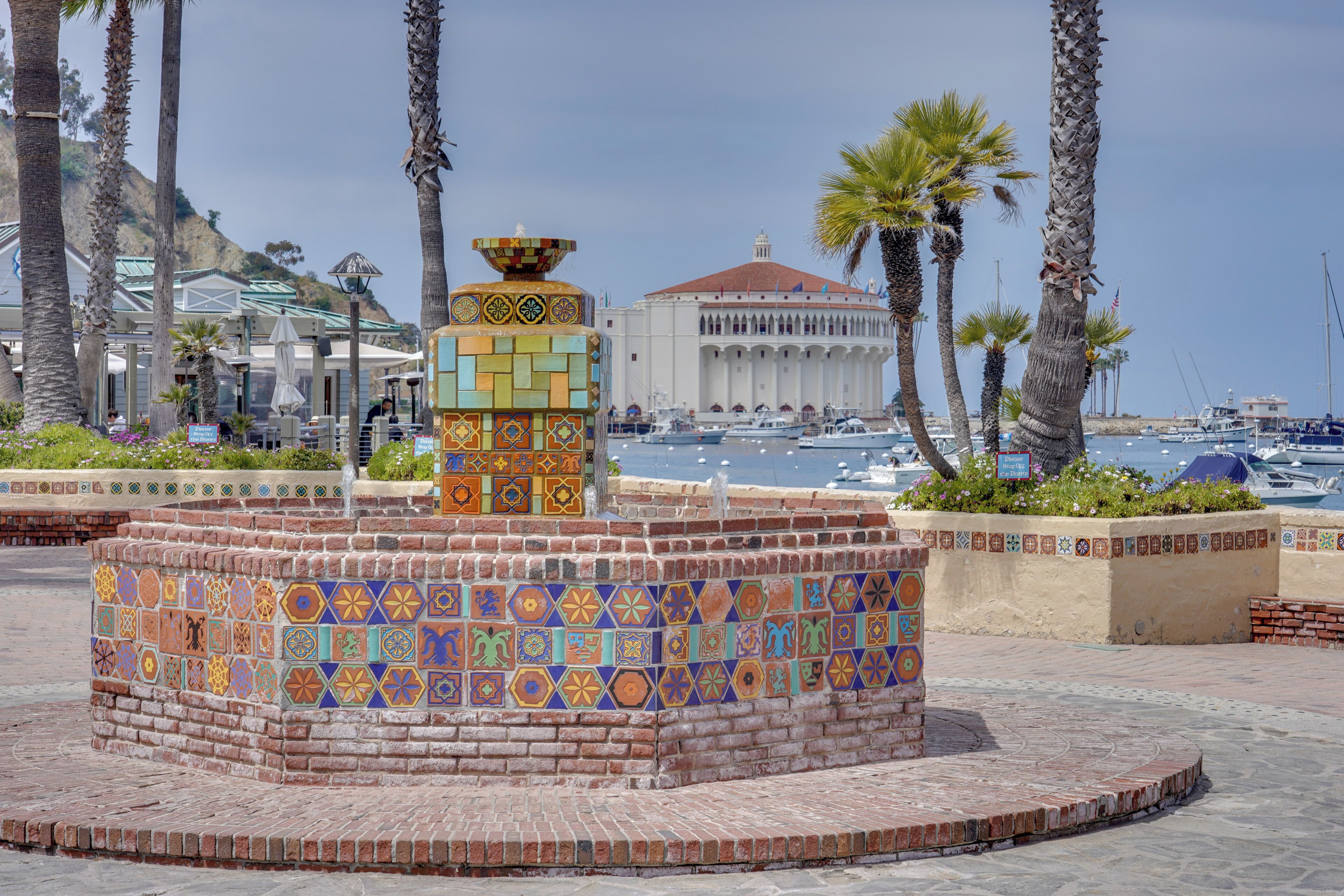 A tiled fountain with a decorative top, surrounded by palm trees and a building with a red roof in the background.