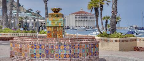 A tiled fountain with a decorative top, surrounded by palm trees and a building with a red roof in the background.