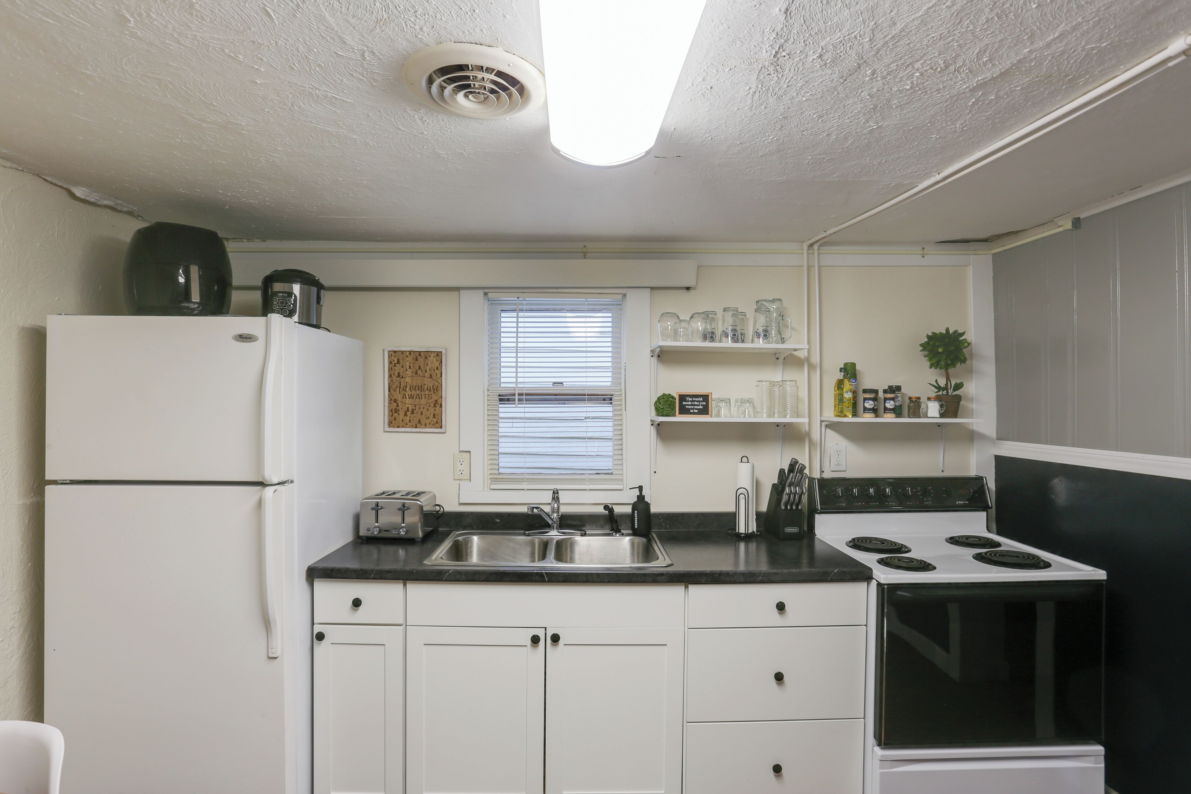 A compact kitchen with white cabinets, a black countertop, a stainless steel sink, a stove, and a window with blinds.
