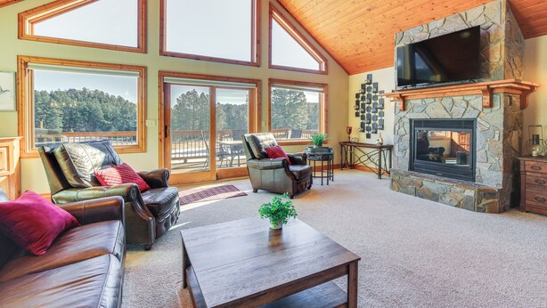 A spacious living room with a stone fireplace, leather sofas, and large windows offering a view of the forest. - Keystone Cabin ~ 9 Mi to Mt Rushmore! (Hermosa)