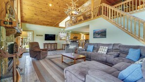 A spacious living area with a large gray sofa, a wooden coffee table, and a fireplace. A wooden staircase with a railing leads to an upper level. - 3-acre Benezette Cabin: Hot Tub, Grill & Mtn View (Benezette)