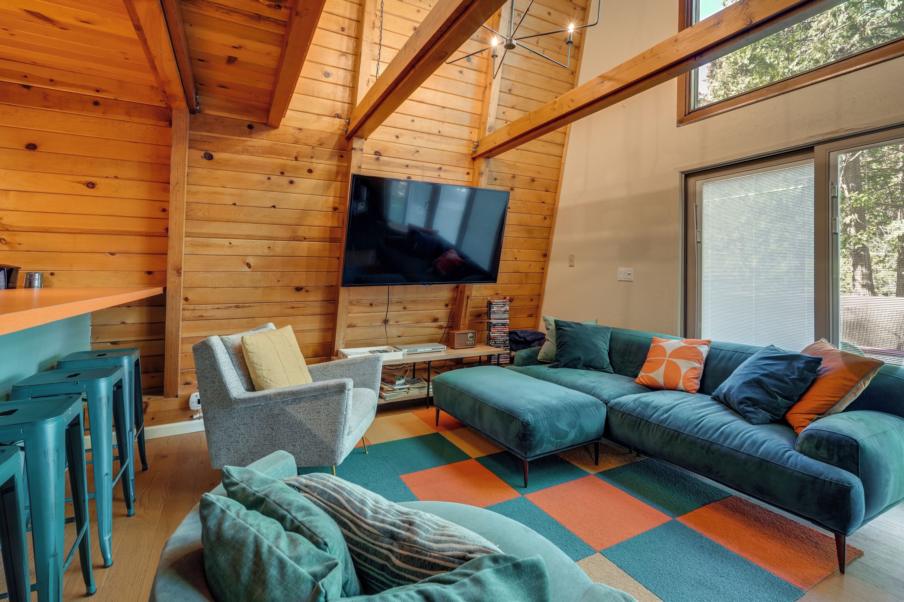 L’intérieur d’un chalet accueillant, avec un plafond en bois, un grand téléviseur fixé au mur, un canapé, un fauteuil et une petite table avec une lampe.