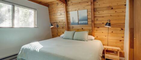 A bedroom with a wooden ceiling, a bed with a light-colored bedspread, two bedside lamps, and a small wooden nightstand.
